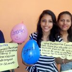 Three young women hold up signs and balloons.