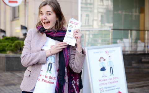 A young woman smiling outside, holding up flyers.